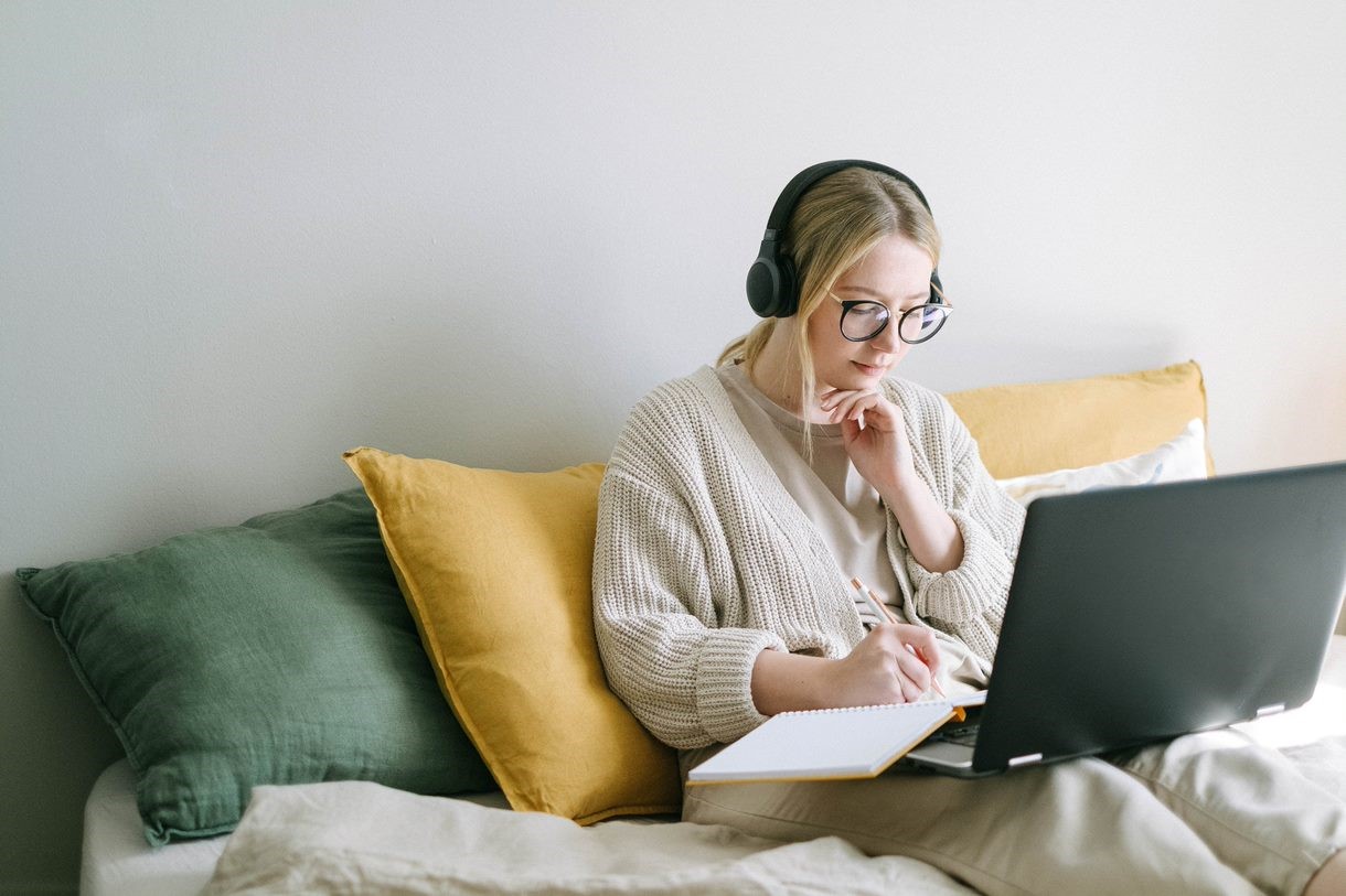woman-sitting-on-bed-with-laptop-in-lap-writing-on-a-notebook